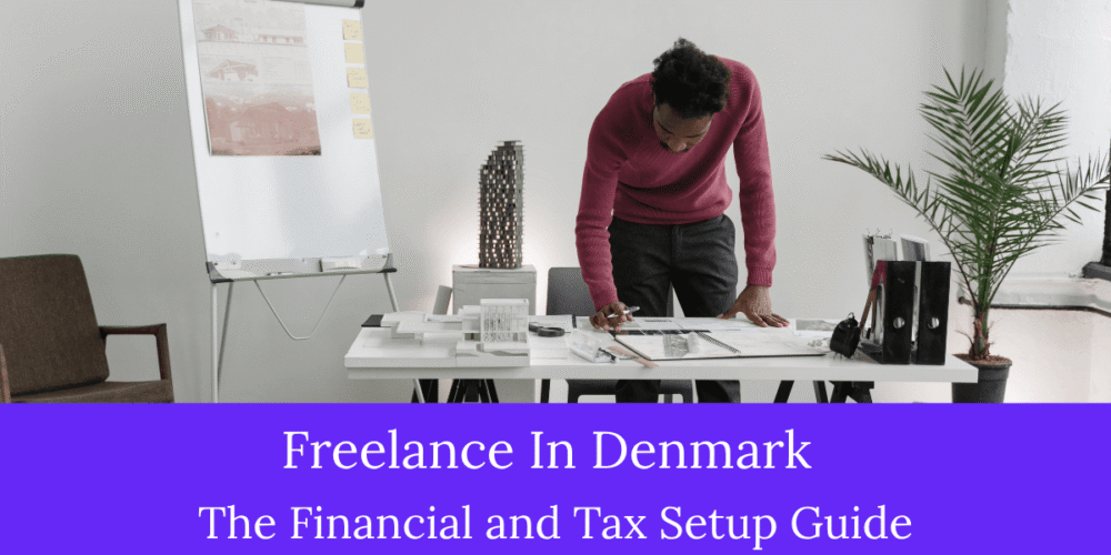 A professional male freelancer in a maroon sweater reviewing documents at a workspace desk with architectural models, featuring text for Freelance In Denmark financial and tax guide.