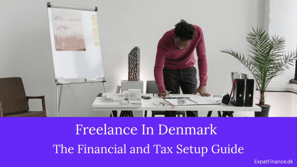 A professional male freelancer in a maroon sweater reviewing documents at a workspace desk with architectural models, featuring text for Freelance In Denmark financial and tax guide.