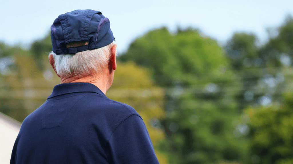 Elderly pensioner in a navy cap and shirt standing outdoors, viewed from behind, with green trees in the background.