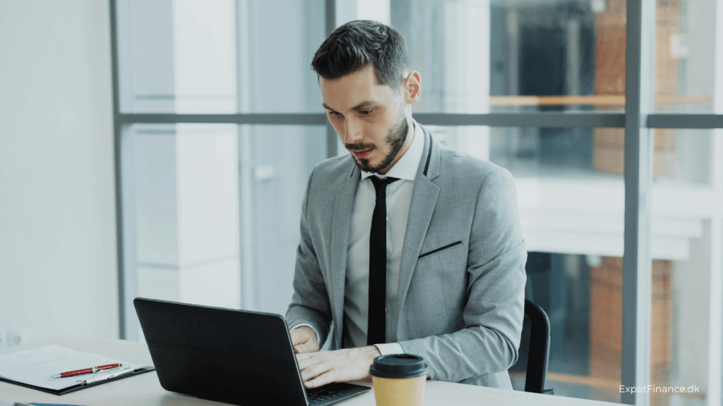 A professional man in a grey suit working on a laptop at an office desk with a coffee cup, representing an expat managing RSU vesting and Danish tax obligations.